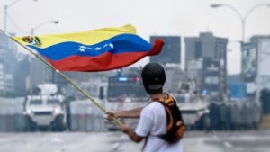 <p>A protestor facing Venezuela’s national guard in May 2017 (image: <a href="https://commons.wikimedia.org/wiki/File:2017_Venezuelan_protests_flag.jpg" target="_blank" rel="noopener">Efecto Eco </a>)</p>