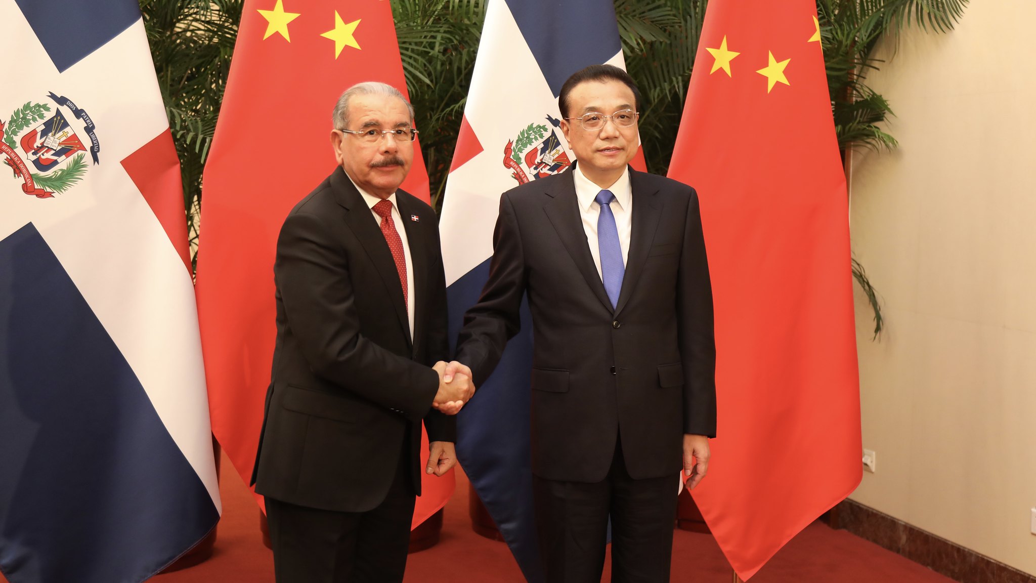 Dois homens de terno e gravata apertam as mãos com as bandeiras da República Dominicana e da China atrás deles.