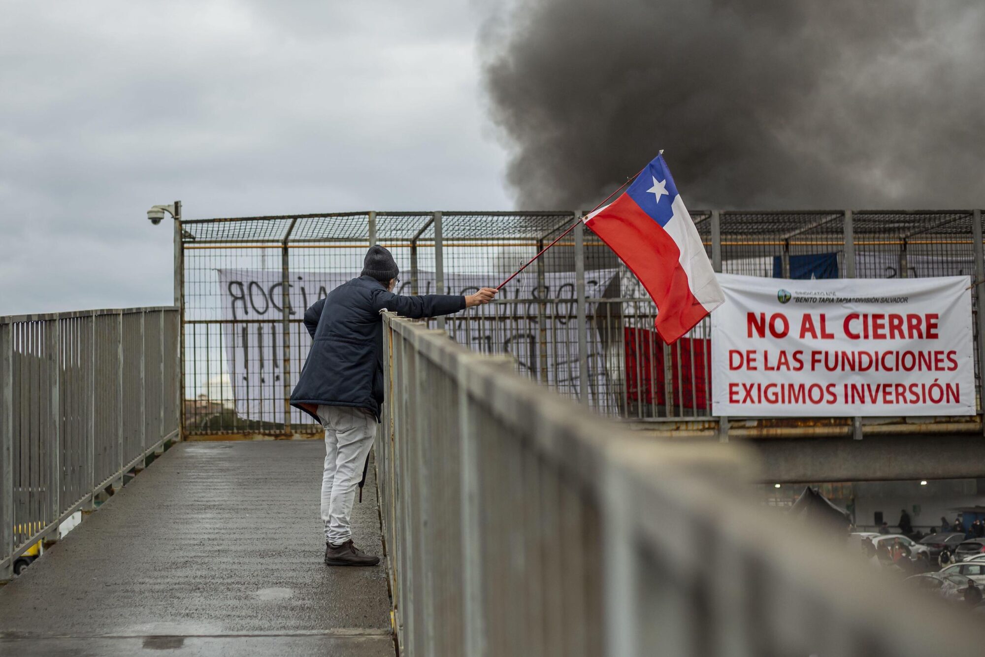 A man protesting the closure of a copper smelting plant waves the Chile flag in front of smoke, standing on a balcony