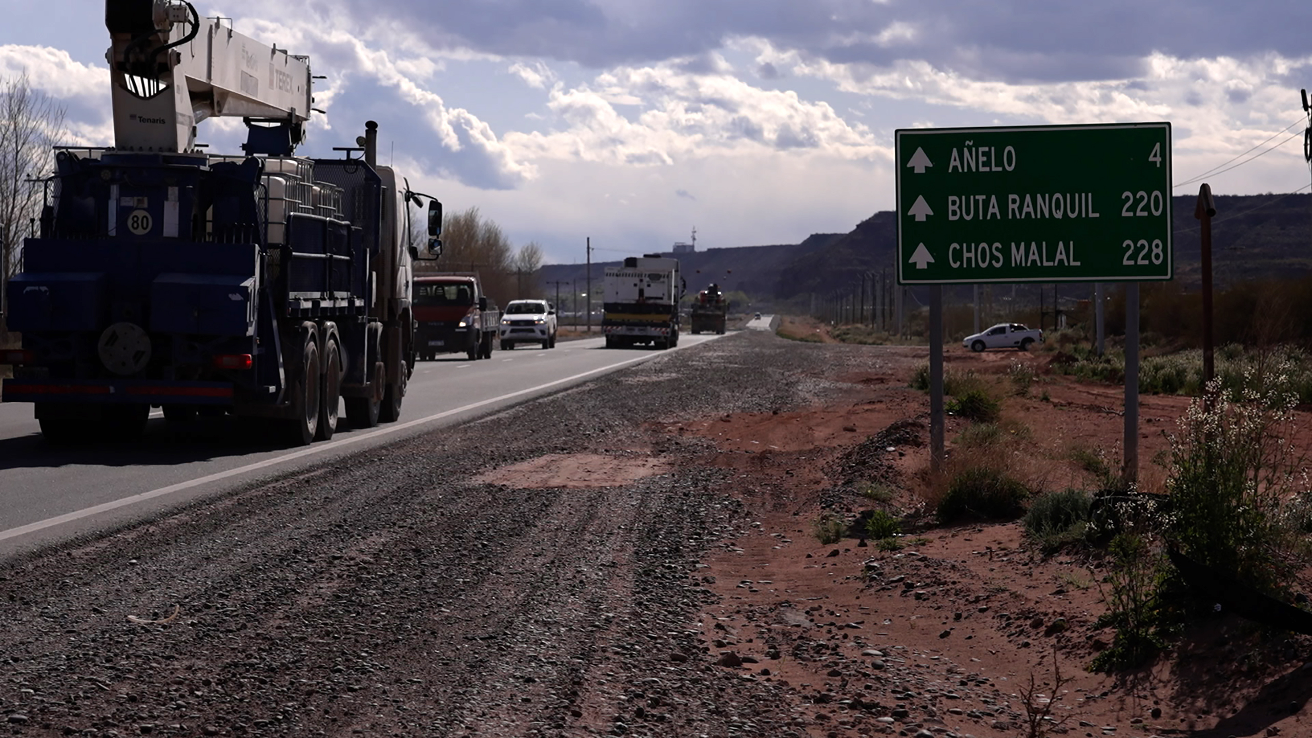 Large trucks facing away drive along a main road to the left, a green road sign to the right shows three places including Anelo, Argentina