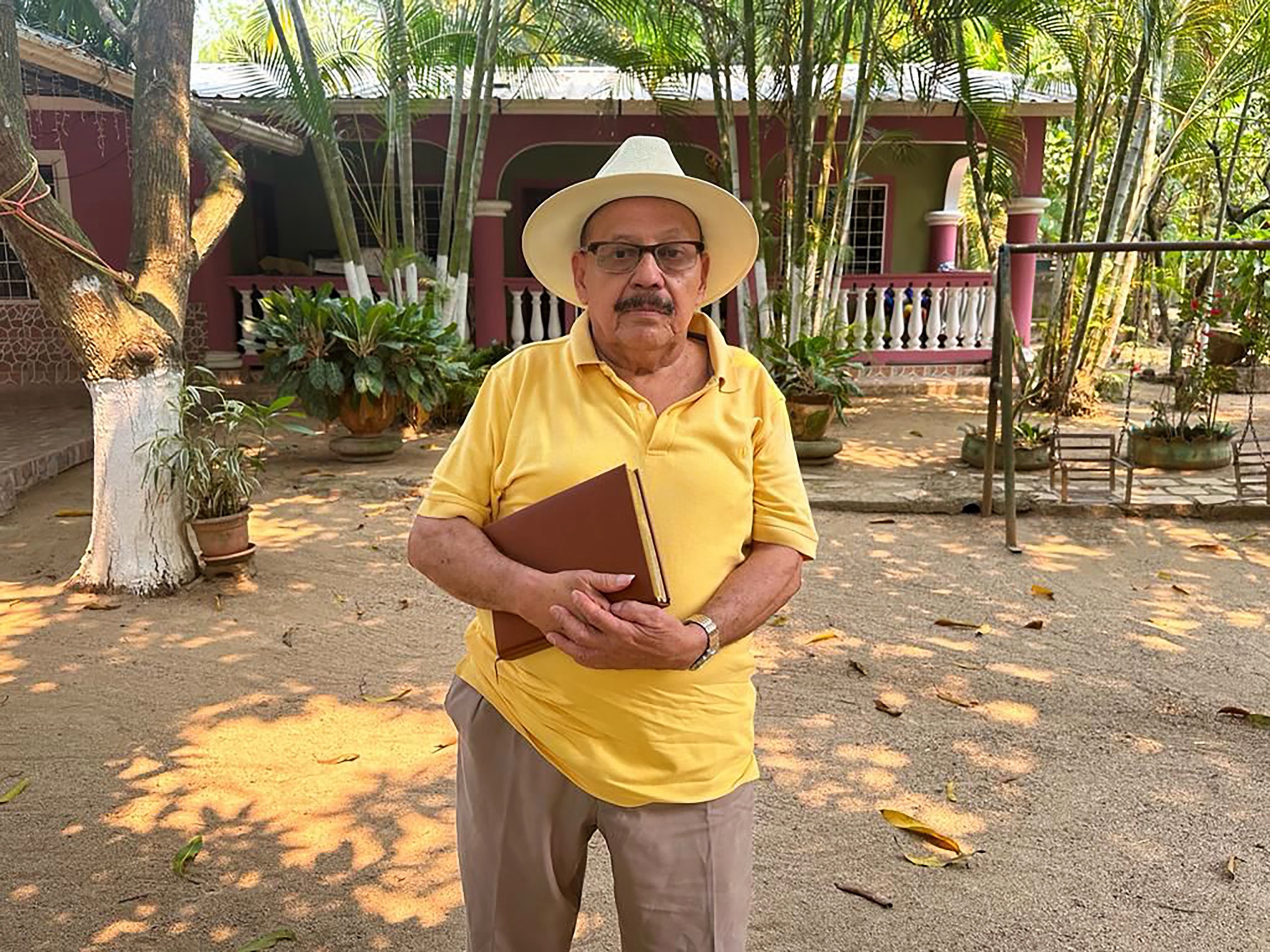 A man in a yellow polo shirt and white hat holding a book, in tropical courtyard