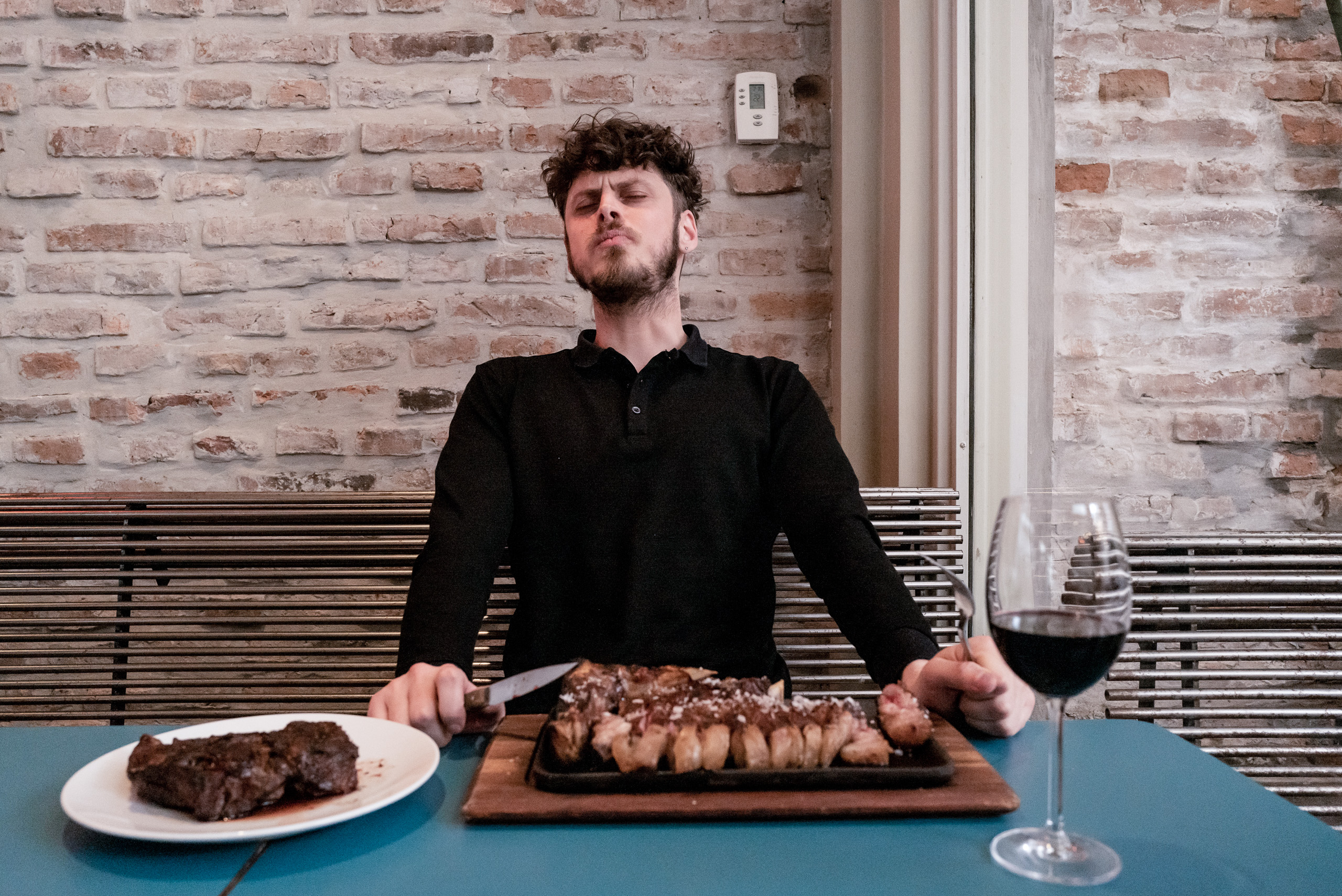 un hombre con una camina negra sentad en una mesa con platos con carne y una copa de vino