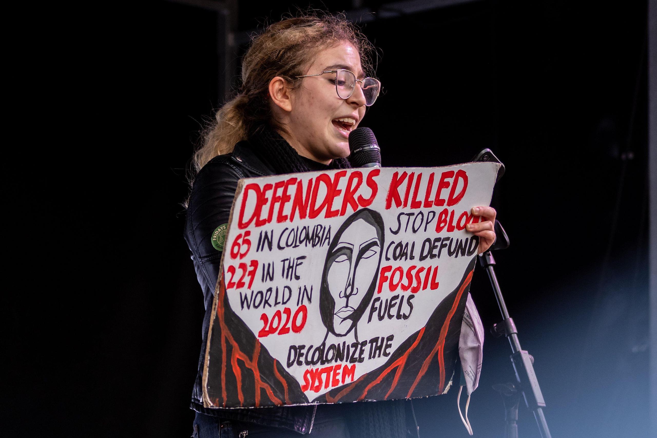 A woman speaks into a microphone, holding a sign that reads: 'Defenders killed, 65 in Colombia, 227 in the world in 2020, stop blood coal, defund fossil fuels, decolonize the system