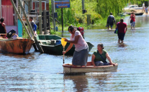 woman paddling small boat on flooded street with people walking in calf deep water in background